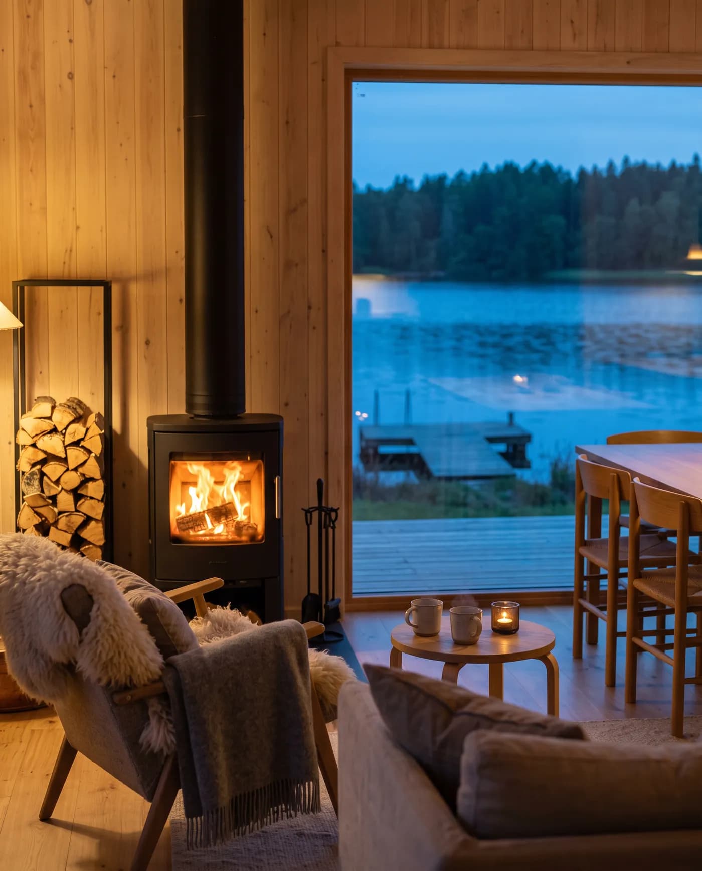 Kaminofen mit Feuer, Sessel mit Fell und Decke, im Fenster der See am Abend